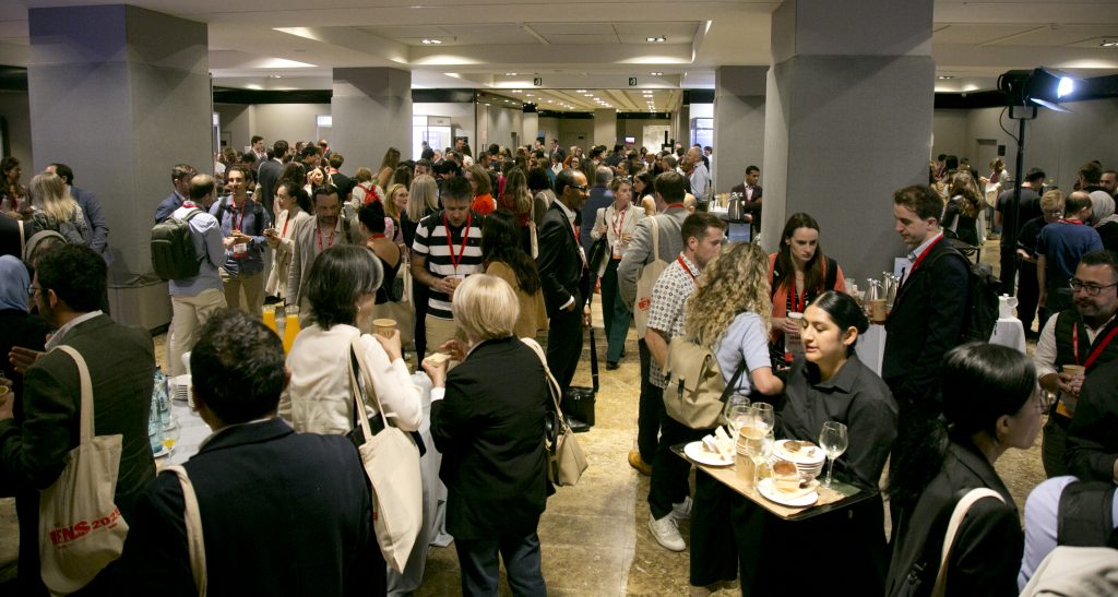 Un nutrido grupo de personas se reúne en un amplio local cubierto en el IFNS 2025 Madrid, muchas de ellas de pie junto a mesas con comida y bebida, socializando y llevando los cordones del evento. Parece tratarse de una conferencia o un evento de networking.