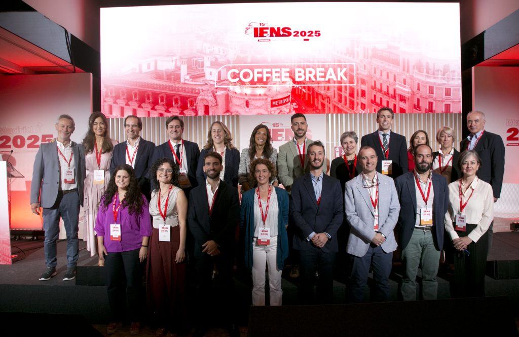 Un grupo de 20 personas posa para una foto en el escenario de una conferencia. El fondo muestra una gran pantalla con el texto IENS 2025 COFFEE BREAK, un paisaje urbano y la marca IFNS 2025 Madrid. Todos llevan insignias de la conferencia.