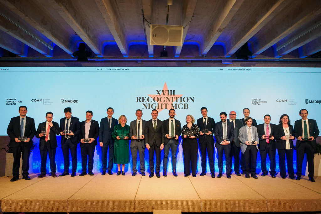 Un grupo de personas en traje de negocios en fila en el escenario sosteniendo premios, con un telón de fondo iluminado en azul que muestra la XVIII Noche de Reconocimiento MCB y los logotipos de los patrocinadores del evento, celebrando embajadores de congresos en Madrid.